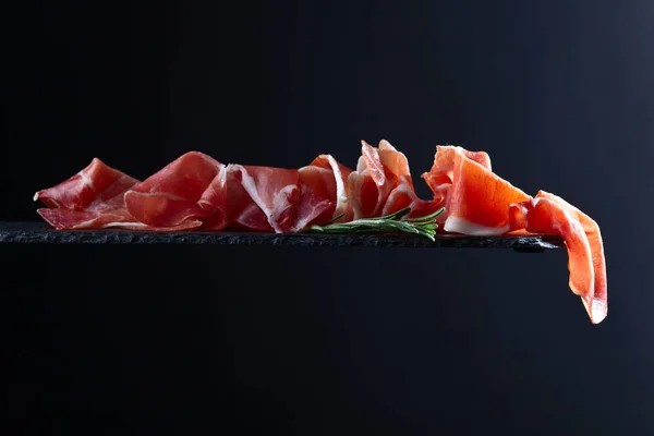 Prosciutto met rozemarijn op een zwarte achtergrond — Stockfoto