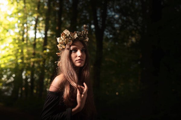 Young beautiful woman in a black dress with a wreath of dry leaves. Dark summer forest.