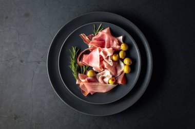 Prosciutto with rosemary and green olives on a black plate. Top 
