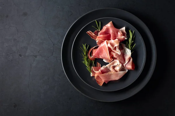 Prosciutto and rosemary on a black plate. Top view. — Stock Photo, Image