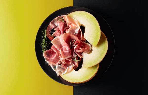 Prosciutto with rosemary and melon on a black plate. — Stock Photo, Image