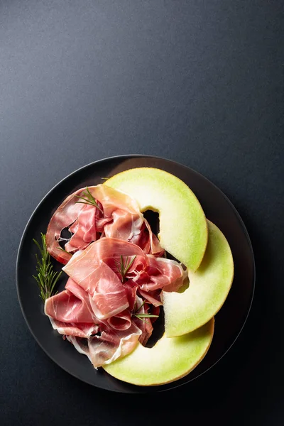 Schinken mit Rosmarin und Melone auf schwarzem Teller. — Stockfoto