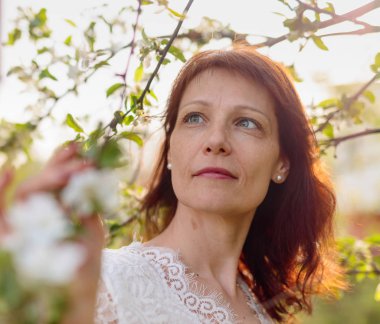 Bahar bahçesinde güzel olgun kadın. Çiçekli bir Elma ağacının yanında beyaz elbiseli çekici bir esmer.