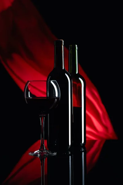Wineglass和一瓶红酒在黑色的反光背景上 红色缎子窗帘在风中飘扬 把重点放在前景上 复制空间 — 图库照片