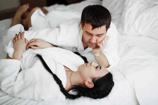 Jovem marido e mulher de casacos brancos brincar e sorrir na cama em um — Fotografia de Stock