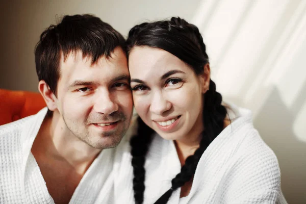 Jovem marido e mulher de casacos brancos brincam e sorriem em uma poltrona laranja em um quarto de hotel. Fechar . — Fotografia de Stock