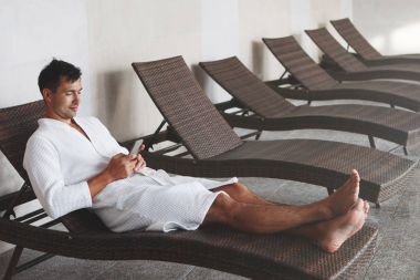 Yakışıklı bir genç adam beyaz terry sabahlık bir yüzme havuzunda bir tembel yatıyor ve smartphone vasıl görünüyor