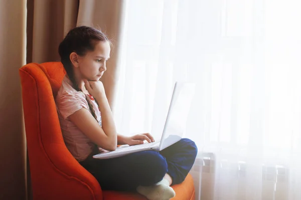 Mooi meisje kind zit op oranje stoel met de laptop. — Stockfoto