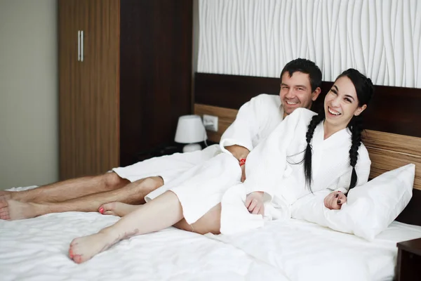 年轻的丈夫和妻子的白色大衣在床上玩耍和微笑在酒店房间里 — 图库照片