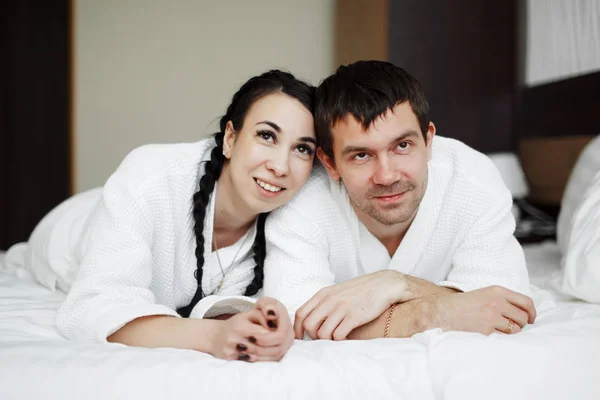 Młody mąż i żona białych płaszczach grać i uśmiech w łóżku w pokoju hotelowym. Z bliska. — Zdjęcie stockowe