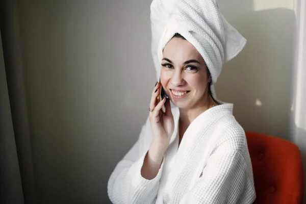Una joven después de una ducha con un teléfono inteligente envuelto en una toalla se sienta en una silla cerca de la ventana — Foto de Stock