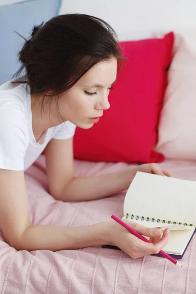 Una chica con una camiseta blanca y jeans está acostada en un sofá con almohadas grises y rojas, sosteniendo un lápiz en la mano y mirando el diario . —  Fotos de Stock