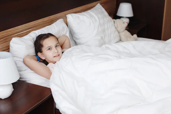 Χαριτωμένος έφηβος κοπέλα βρίσκεται στο κρεβάτι με λευκά ρούχα και φαίνεται στην κάμερα — Φωτογραφία Αρχείου