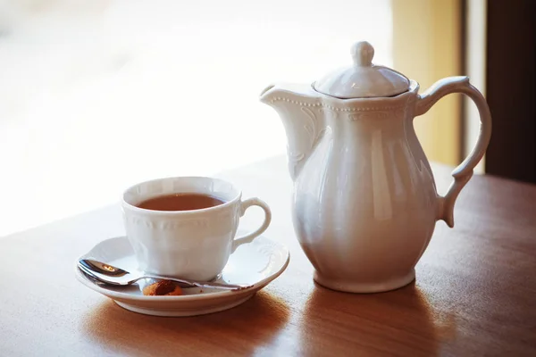 Uma chaleira branca e uma xícara com um pires e uma colher de chá estão sobre a mesa — Fotografia de Stock