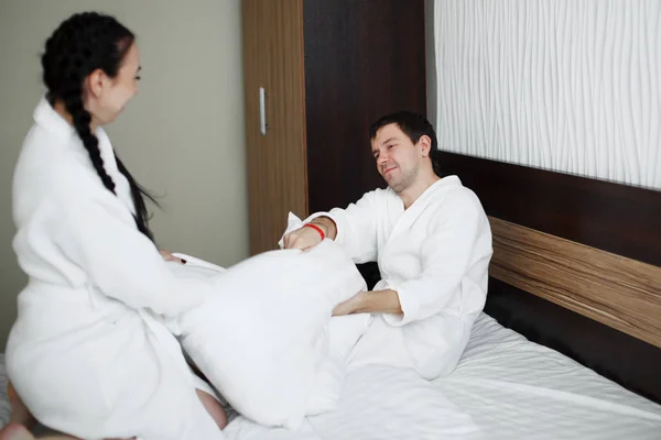 Jovem marido e mulher de casacos brancos brincam e sorriem na cama em um quarto de hotel — Fotografia de Stock