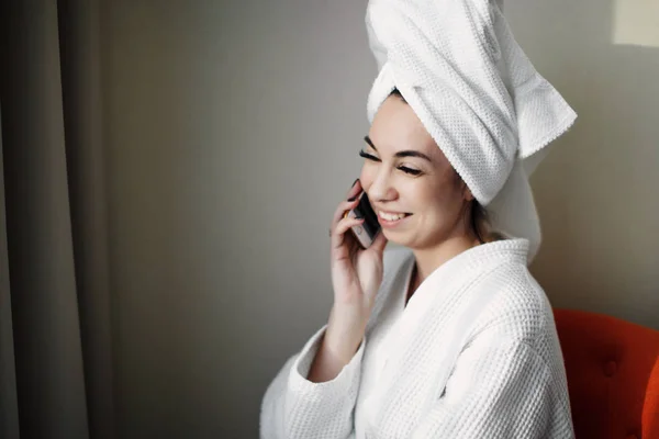 Uma jovem após um banho com um smartphone embrulhado em uma toalha senta-se em uma cadeira perto da janela — Fotografia de Stock
