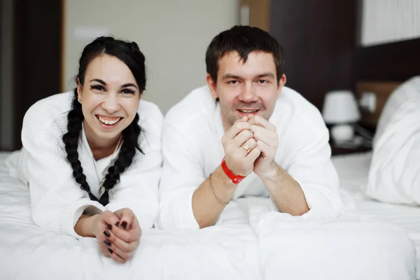 Młody mąż i żona białych płaszczach grać i uśmiech w łóżku w pokoju hotelowym. Z bliska. — Zdjęcie stockowe