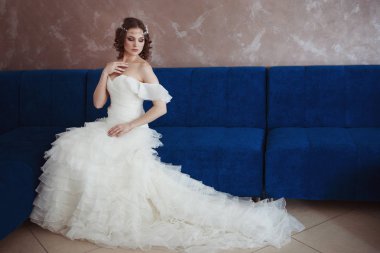 Sevimli genç gelin saçında bir diadem ile bir beyaz gelinlik kapalı mavi kanepede oturuyor