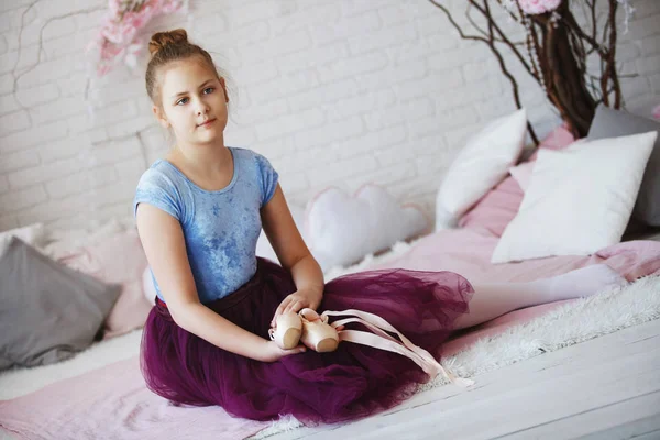 Uma jovem bailarina em roupas azuis e uma saia roxa com pointes rosa senta-se em uma cama branca com travesseiros . — Fotografia de Stock