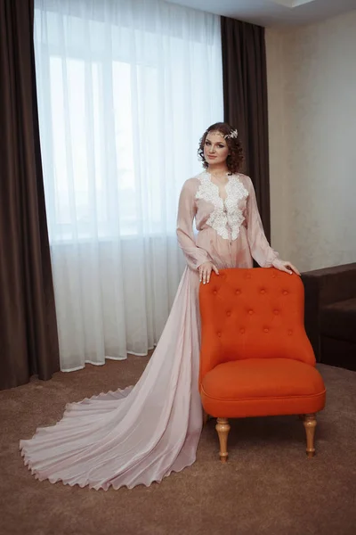 Cute young bride in a wedding white dress with a diadem in her hair stay with orange armchair indoors — Stock Photo, Image