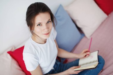 Bir kız beyaz bir T-shirt ve kot bağdaş kurup bir kanepe ile gri yastık oturur ve kırmızı günlüğü görünüyor.