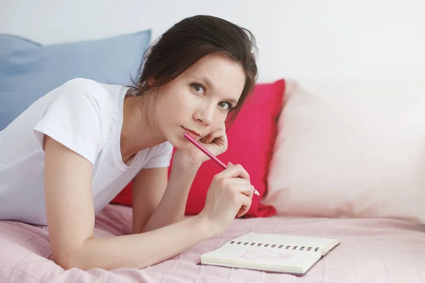 Una chica con una camiseta blanca y jeans está acostada en un sofá con almohadas grises y rojas, sosteniendo un lápiz en la mano y mirando el diario . — Foto de Stock
