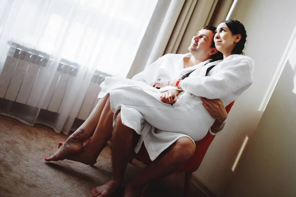 年轻的丈夫和妻子的白色大衣在床上玩耍和微笑在酒店房间里. — 图库照片