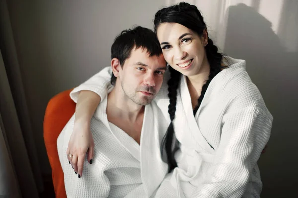 年轻的丈夫和妻子的白色大衣在一个橙色的扶手椅在酒店房间里玩耍和微笑。特写. — 图库照片