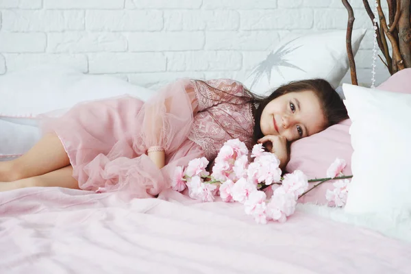 Menina em um vestido rosa encontra-se em uma cama com almofadas brancas e flores — Fotografia de Stock