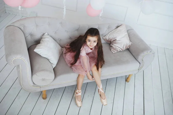 A young ballerina in a pink dress is sitting on a gray sofa with pointes in hands — Stock Photo, Image