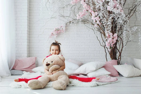 Uma jovem menina de cabelos longos em uma camiseta rosa e shorts e golfs mentiras, abraços com um urso de brinquedo na cama com almofadas e galhos de árvore — Fotografia de Stock