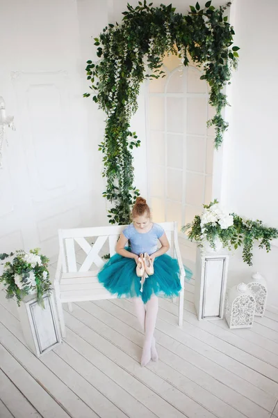 Uma jovem bailarina em uma saia azul com pointes rosa senta-se em um banco de madeira branca . — Fotografia de Stock