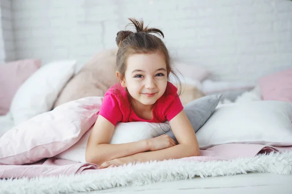 A young long-haired girl in a pink T-shirt lying in bed with pillows cheerfully and slyly looks at the camera