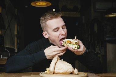 Genç yakışıklı sarışın adam kafede bir sandviçi ısırıyor..