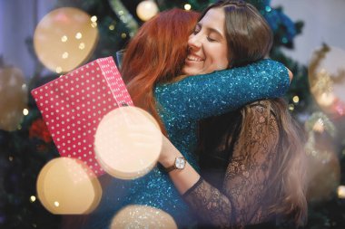 İki kız arkadaş Noel ağacının yanında birbirlerine hediye kutuları veriyorlar..