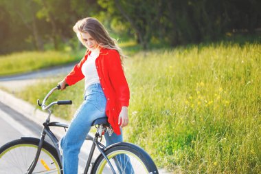 Şehir parkında bisiklet süren bir kadın. Uzun saç, kot pantolon, kırmızı ve beyaz gömlek..