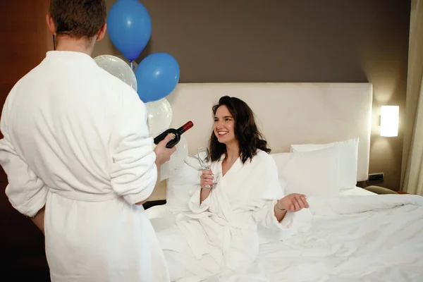 两个情人 一个男人提供一个女人在床上喝酒 酒店房间 白色浴衣 节日生日 — 图库照片