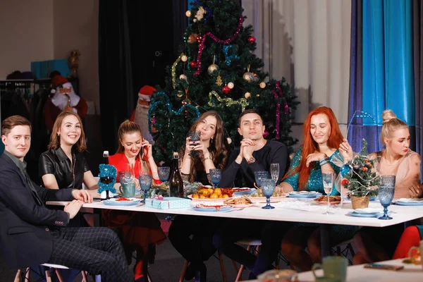 一群快乐的朋友在圣诞树旁的餐桌旁 用花环庆祝新年 女人和男人笑 用香槟酒杯庆祝 — 图库照片