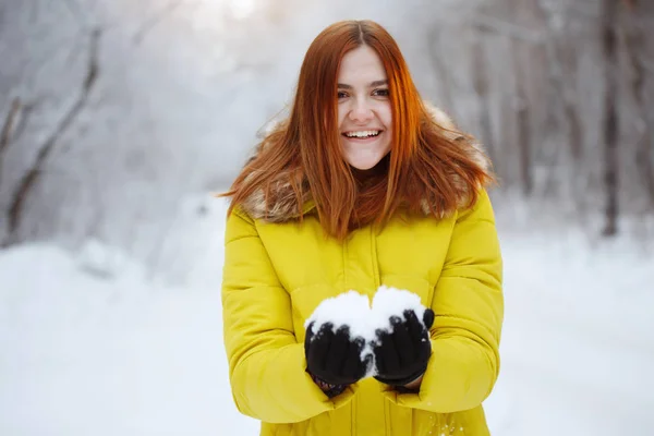 マスタードジャケットの若い かなり陽気な赤い髪の長い髪の女性は 雪の森の中で外の冬に彼女の手から雪片を投げ 笑っています — ストック写真