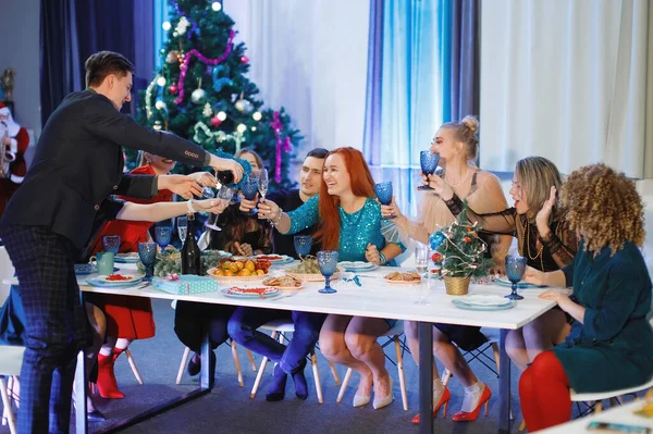 一群快乐的朋友庆祝圣诞节或新年 一个男人往杯子里倒香槟 女人笑着坐在圣诞树旁的桌子旁 — 图库照片