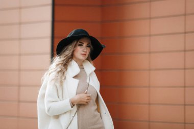 Portrait of a blonde lady in a black hat outdoors, long hair, beige coat.