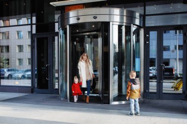 İki çocuklu bir anne, cam ve çelikten yapılmış bir ofis binası ya da otelin girişinde, gökyüzünün yansıması ve döner bir kapıyla..