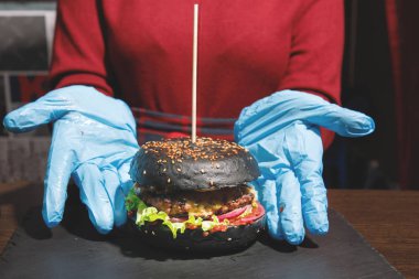 Aşçı ellerinde kırmızı önlüklü ve mavi eldivenli bir hamburger sunuyor..