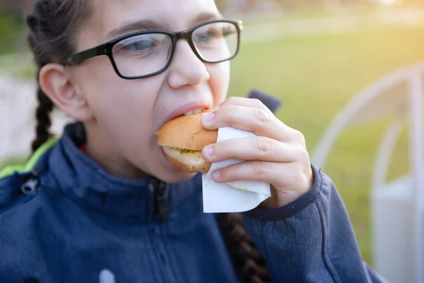 Teen girl eats a hamburger on a bench in the city quarter. Harmful food, fight obesity.