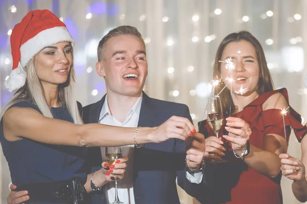 Group of people are standing with glasses of champagne and sparklers on a holiday in caps and laughing. Selected, soft focus. Vintage effect of old film. Filter for noise and grain.