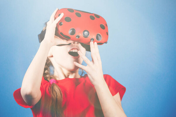 Blondy teen girl plays with virtual reality glasses. Computer games, blue background, emotions.