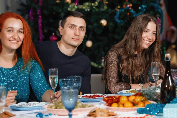 友達の陽気な会社は ガーランドとクリスマスツリーの近くのテーブルで新年を祝います 女性と男性は笑いシャンパングラスで喜びます — ストック写真