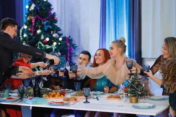 一群快乐的朋友庆祝圣诞节或新年 一个男人往杯子里倒香槟 女人笑着坐在圣诞树旁的桌子旁 — 图库照片