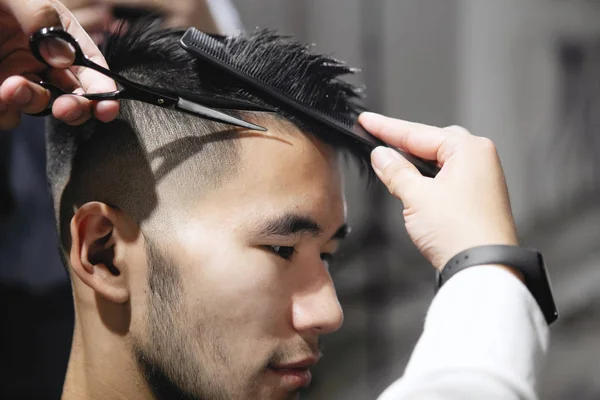 Young guy hairdresser cuts hair with scissors to a man in a beauty salon.