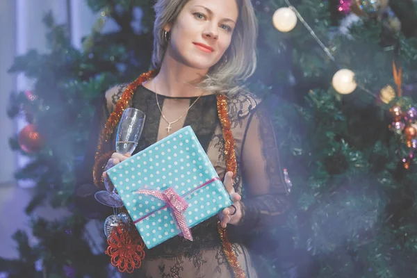 Chic woman rejoices with a gift box near a Christmas tree. A woman laughs, smiles, poses. Special vintage noise and grain filter, blurry lights.
